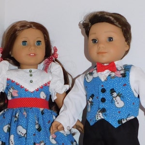 May include: Two dolls dressed in blue and red winter outfits with snowman patterns. The girl doll has long brown hair and is wearing a blue dress with a red belt. The boy doll has short brown hair and is wearing a blue vest with a red bow tie.
