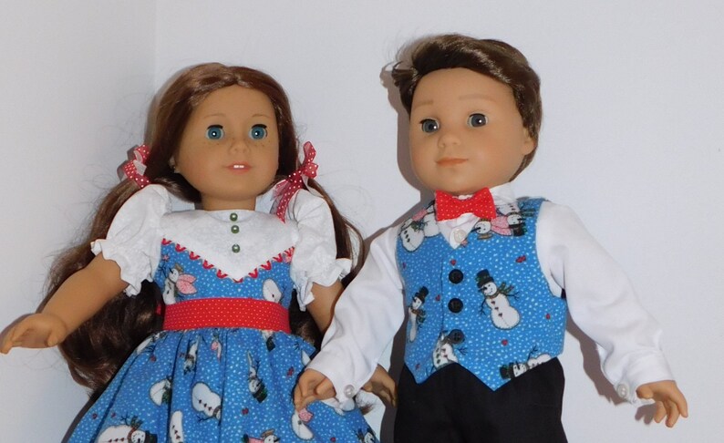 May include: Two dolls dressed in blue and red winter outfits with snowman patterns. The girl doll has long brown hair and is wearing a blue dress with a red belt. The boy doll has short brown hair and is wearing a blue vest with a red bow tie.