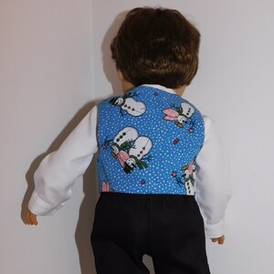 May include: A blue vest with a snowman pattern for a doll. The vest has a white background with red hearts and a blue background with white snow. The vest is worn over a white shirt and black pants.