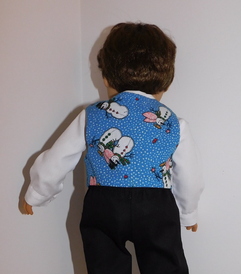 May include: A blue vest with a snowman pattern for a doll. The vest has a white background with red hearts and a blue background with white snow. The vest is worn over a white shirt and black pants.