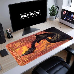 Puede incluir: Alfombrilla de ratón rectangular con una ilustración de un dragón negro sobre llamas naranjas y amarillas. La alfombrilla tiene un borde rojo con texto. El monitor muestra el texto "MUFTPADS".