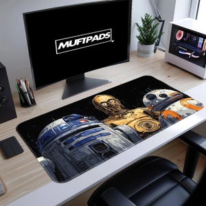 May include: A black rectangular desk mat featuring a Star Wars design with R2-D2, C-3PO, and BB-8. The mat is on a white desk with a computer monitor displaying the text "MUFTPADS". A small potted plant and computer tower are also visible.