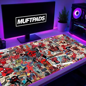 May include: A large rectangular desk mat featuring a collage of Spider-Man comic book panels. The mat is surrounded by a glowing purple light. The computer monitor displays the text "MUFTPADS". A computer tower with colorful lights is visible.