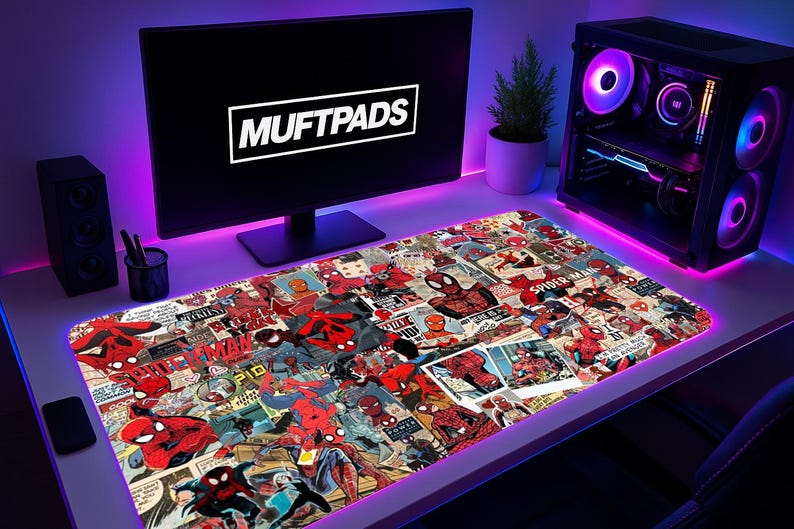 May include: A large rectangular desk mat featuring a collage of Spider-Man comic book panels. The mat is surrounded by a glowing purple light. The computer monitor displays the text "MUFTPADS". A computer tower with colorful lights is visible.