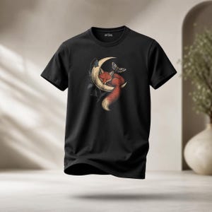 Könnte beinhalten: Ein schwarzes Kurzarm-T-Shirt mit einem Grafikdruck eines roten Fuchses, der zusammengerollt auf einem cremefarbenen Halbmond schläft. Das Design zeigt einen Nachtfalter über dem Fuchs und blaue glockenförmige Blumen am Rand des Mondes.