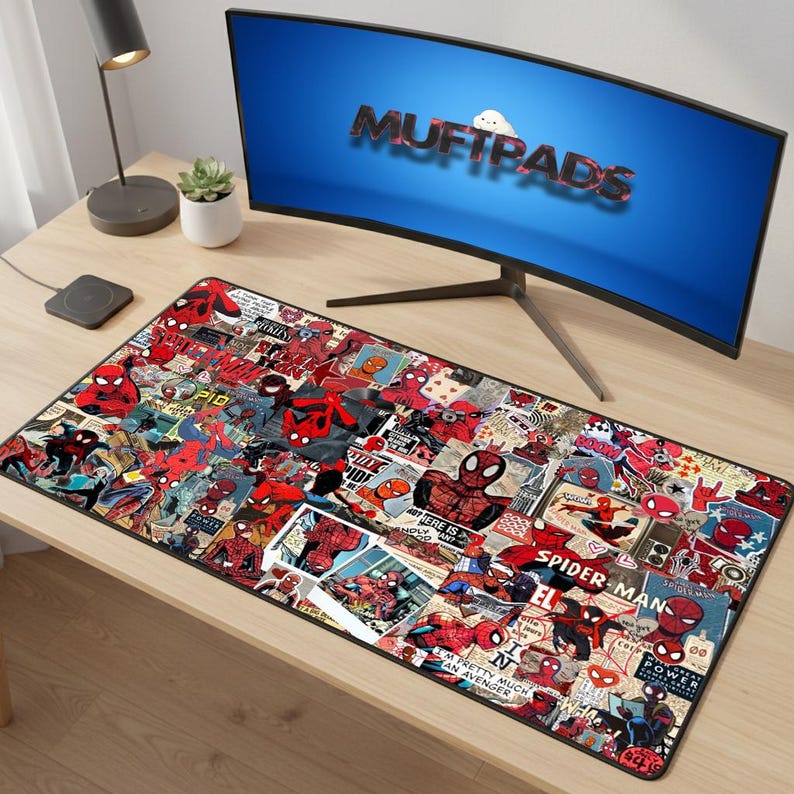 May include: A rectangular desk mat featuring a collage of Spider-Man comic book images in red, black, and blue. The mat is placed on a light-colored wooden desk, with a computer monitor displaying the text "MUFTPADS".