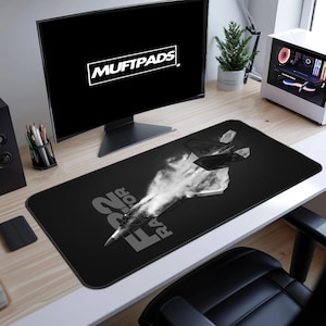 Könnte beinhalten: Schwarze rechteckige Schreibtischunterlage mit einem Graustufenbild eines Kampfflugzeugs mit Kondensstreifen. Der Text "F22 RAPTOR" ist weiß auf der Matte aufgedruckt. Die Marke "MUFTPADS" wird auf dem Computermonitor angezeigt.