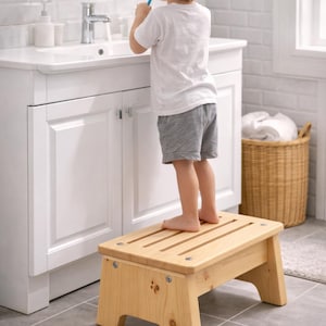 Op de afbeelding: Een lichtgekleurde houten opstap met een lattenbodem. De kruk staat in een badkamer, met een kind dat erop staat en zijn tanden poetst bij de wastafel. De kruk is ongeveer 30 cm hoog.
