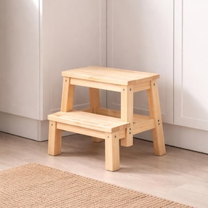 Puede incluir: Un taburete de madera de dos escalones con acabado natural. El taburete tiene una parte superior rectangular y un escalón inferior, sostenidos por patas robustas. La veta de la madera es visible, y el taburete está colocado sobre un suelo claro con una alfombra tejida delante.