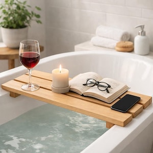 May include: A wooden bathtub tray holds a lit candle, an open book with black-framed glasses, and a smartphone. A glass of red wine sits on the tray, with a bathtub filled with water in the foreground. The scene suggests relaxation and self-care.