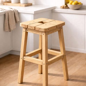 Op de afbeelding: Een vierkante houten kruk met vier poten en een zitting van latten met een vierkante uitsparing in het midden. De kruk is gemaakt van lichtgekleurd hout met een zichtbare nerf en is gemonteerd met metalen bevestigingen bij de verbindingen.