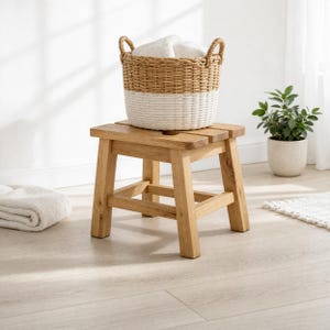 May include: A natural wood stool with a woven basket. The basket is two-toned, with a natural brown top and a white bottom. The basket contains folded white towels. A small potted plant is in the background.