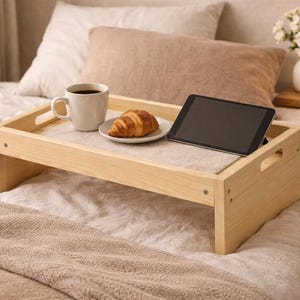 May include: A wooden bed tray holds a cup of coffee, a croissant on a plate, and a tablet. The tray is on a bed with white and beige bedding. A vase of white flowers and a lamp are on a nightstand.