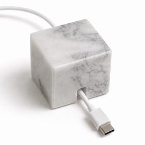 Puede incluir: Un cubo de mármol blanco con vetas grises, forma cuadrada y acabado liso. Un cable de carga blanco con conector USB-C sale de una pequeña ranura vertical cortada en la cara frontal del bloque de piedra, funcionando como una base de soporte.