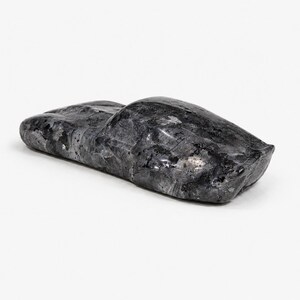 May include: A polished, dark gray stone sculpture carved into the abstract, elongated shape of a vehicle. The surface features natural mineral inclusions and crystalline patterns that create a mottled, reflective appearance across the entire form.