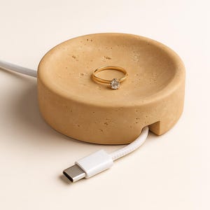 May include: A tan-coloured, circular ring holder with a gold ring featuring a clear stone. A white USB-C cable is inserted into the side. The ring holder is made of a textured material.