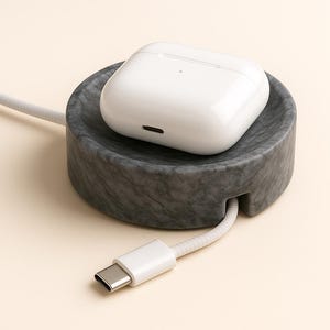 May include: A white wireless earbud charging case sits on a grey marble charging station. A white charging cable with a USB-C connector is plugged into the station. The station is round with a cable slot.