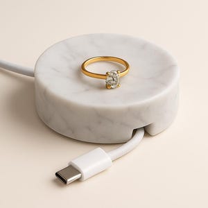 Puede incluir: Un anillo de oro con una gema ovalada transparente descansa sobre una base de carga de mármol blanco. Un cable USB-C blanco está conectado a la base. El anillo es el punto focal.
