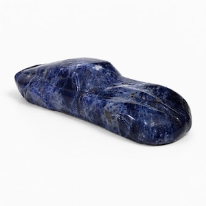 May include: A polished, deep blue sodalite stone carved into the smooth, streamlined shape of a sports car. The mineral features natural white and dark blue marbling throughout its surface, creating a glossy, textured appearance in this sculpture.