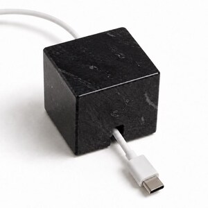 May include: A black marble cube cable holder with a smooth, polished finish and natural grey veining. A white charging cable with a USB-C connector is threaded through a small square notch cut into the bottom front edge of the stone block.