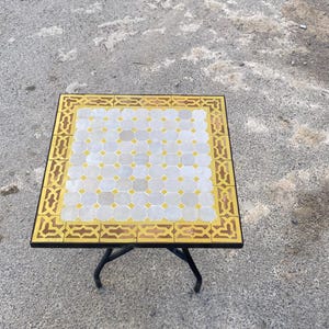 May include: Square mosaic tile table with a black metal frame and legs. The tabletop features a pattern of white and yellow tiles, framed by a gold border. The table is approximately 24 inches wide and 24 inches long.