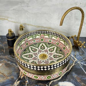 Puede incluir: Un lavabo de cerámica decorativo con un detallado estampado floral en verde, rosa y blanco. El lavabo tiene un borde dorado y se combina con un grifo dorado. Se ven dos dispensadores de jabón.