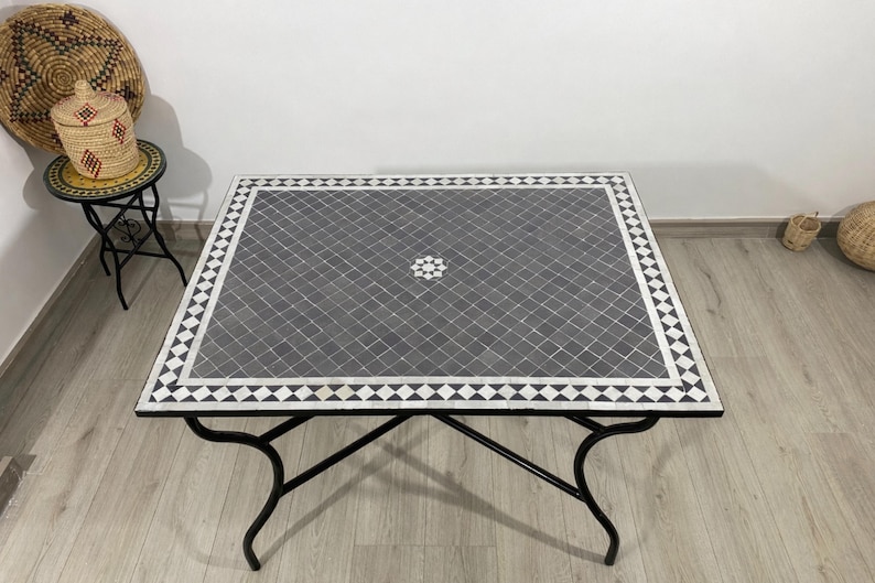 K&ouml;nnte beinhalten: Quadratischer Mosaiktisch mit schwarzem Metallgestell. Die Tischplatte hat ein grau-wei&szlig;es geometrisches Muster mit einem zentralen Blumendesign. Der Tisch ist etwa 91 cm gro&szlig;.
