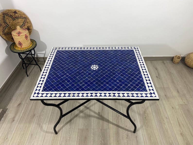 K&ouml;nnte beinhalten: Quadratischer Couchtisch mit blauer Mosaikfliesenplatte und schwarzem Metallgestell. Das Mosaik hat einen wei&szlig;en Rand und ein zentrales Blumendesign. Die Tischma&szlig;e betragen ca. 91 cm x 91 cm.