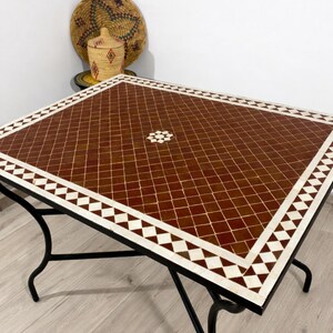 K&ouml;nnte beinhalten: Quadratischer Mosaiktisch mit braunem und wei&szlig;em geometrischem Muster. Der Tisch hat ein schwarzes Metallgestell und Beine. Dekorative gewebte K&ouml;rbe sind im Hintergrund.