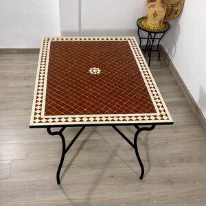 K&ouml;nnte beinhalten: Rechteckiger Couchtisch mit einer geometrischen Fliesenplatte in Braun und Wei&szlig;. Der Tisch hat ein schwarzes Metallgestell mit gebogenen Beinen. Die Tischplatte weist ein Rautenmuster mit einem zentralen Blumendesign auf. Der Tisch ist ca. 122 cm lang und 61 cm breit.