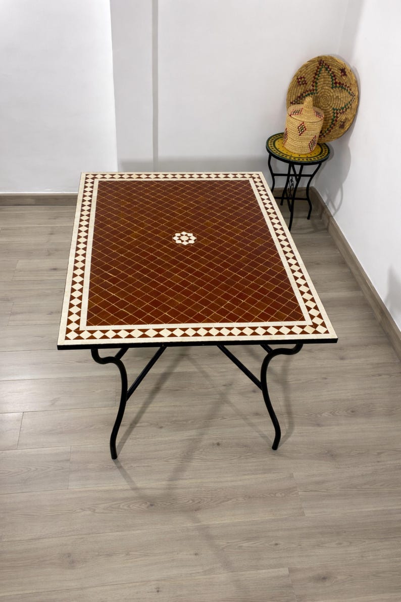 K&ouml;nnte beinhalten: Rechteckiger Couchtisch mit einer geometrischen Fliesenplatte in Braun und Wei&szlig;. Der Tisch hat ein schwarzes Metallgestell mit gebogenen Beinen. Die Tischplatte weist ein Rautenmuster mit einem zentralen Blumendesign auf. Der Tisch ist ca. 122 cm lang und 61 cm breit.