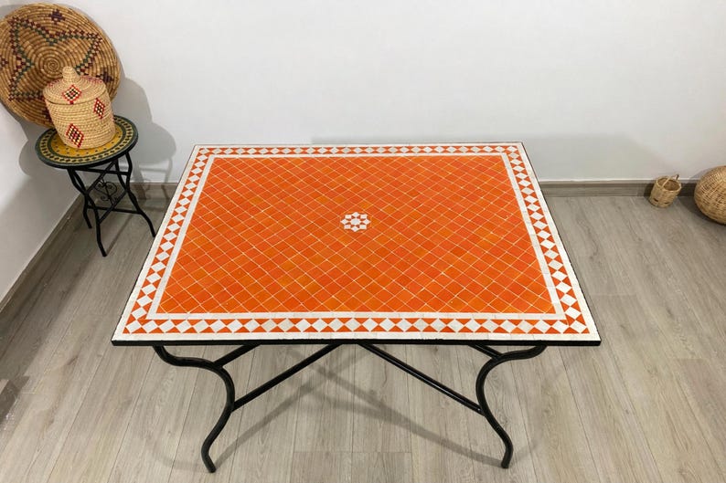 K&ouml;nnte beinhalten: Orangener Mosaiktisch mit wei&szlig;em geometrischen Rand und schwarzem Metallgestell. Der rechteckige Tisch hat ein dekoratives Blumendesign in der Mitte. Der Tisch ist ca. 100 cm lang und 75 cm breit.