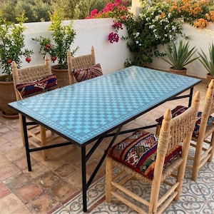 May include: A rectangular outdoor table with a blue and white checkered pattern top, supported by black metal legs. Woven chairs with patterned cushions surround the table. Potted plants and flowers are in the background.