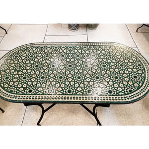 May include: Oval-shaped outdoor table with a green and white mosaic tile top. The intricate pattern features geometric shapes and floral motifs. The table has a black metal base and measures approximately 60 inches long.