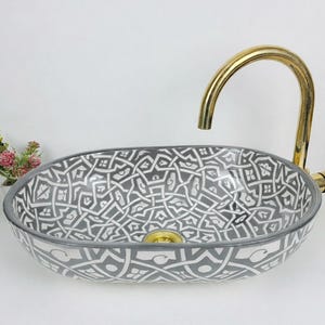 May include: Oval ceramic bathroom sink with a grey and white geometric pattern. A gold-coloured tap is installed on the side. The sink is on a white surface. A small bouquet of flowers is on the left.