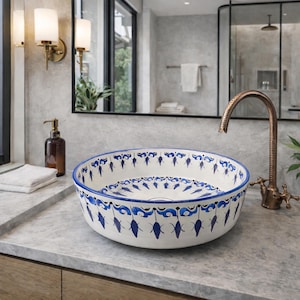 May include: A white ceramic vessel sink with a blue decorative pattern. The sink is round and sits on a gray countertop. A bronze faucet and wall sconces are visible in the background. The sink is a bathroom fixture.