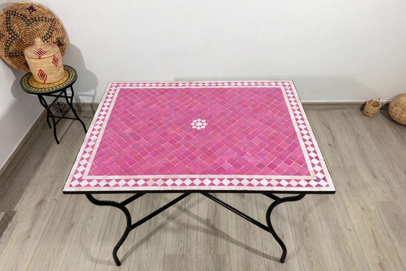 K&ouml;nnte beinhalten: Quadratischer Couchtisch mit Mosaikfliesen in Rosa- und Wei&szlig;t&ouml;nen. Der Tisch hat ein schwarzes Metallgestell mit geschwungenen Beinen. Das Mosaikdesign zeigt ein zentrales Blumenmuster und einen geometrischen Rand.