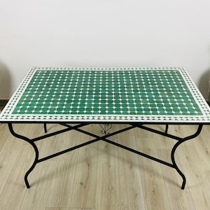 May include: A rectangular coffee table with a green and white tiled top. The table has a black wrought iron base with curved legs. The tabletop is decorated with a repeating pattern of green and white tiles. The table is a decorative piece for a living room.