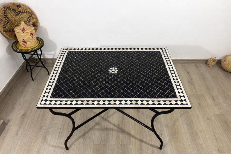 K&ouml;nnte beinhalten: Quadratischer Tisch mit schwarzen Mosaikfliesen, wei&szlig;em geometrischen Rand und zentralem Blumendesign. Der Tisch hat ein schwarzes Metallgestell und misst ca. 99 cm im Quadrat.