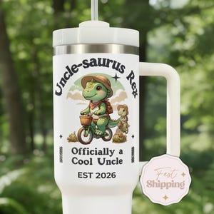 May include: White insulated tumbler with a handle, featuring a green dinosaur riding a bicycle with the text "Uncle-saurus Rex" and "Officially a Cool Uncle EST 2026". The design includes a smaller dinosaur and decorative elements.