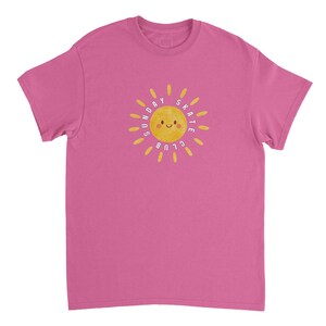 Könnte beinhalten: Magentafarbenes T-Shirt mit einem gelben Sonnensymbol und dem Text "SUNDAY SKATE CLUB". Die Sonne hat ein lächelndes Gesicht und ist von gelben Strahlen umgeben. Das Shirt besteht aus einem weichen Material.