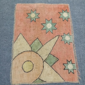 May include: A rectangular rug featuring a muted coral background with a large, stylized geometric sun motif in tan and green in the lower left corner. The upper portion is decorated with five eight-pointed green and tan stars. The edges have fringe.