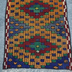 May include: A rectangular, hand-woven rug with a vibrant geometric pattern. The rug features a central diamond design in green, yellow, and red, surrounded by a repeating pattern of blue, orange, and red rectangles. White fringe edges the rug.
