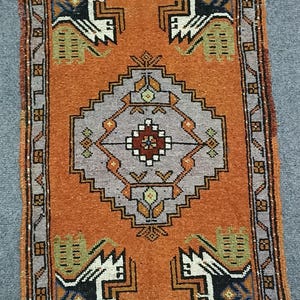May include: An orange and gray rectangular rug with a geometric pattern. The rug features a central diamond shape and repeating stylized animal figures in black, white, and green. The border is a darker shade of orange.