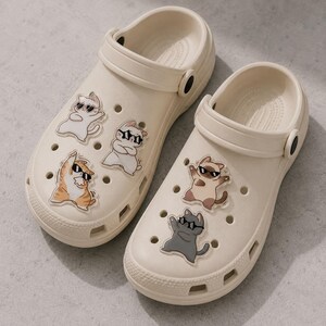 May include: A pair of cream-colored clogs adorned with cartoon cat decals wearing sunglasses. The cats are in various poses, adding a playful touch to the footwear. The clogs have a classic design with ventilation holes.
