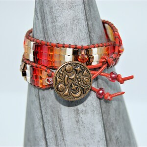 May include: Wrap bracelet with a mix of orange, red, and white beads. The bracelet has a leather strap and a decorative bronze button clasp. The bracelet is wrapped around a gray wooden form.