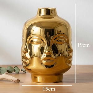 May include: A gold-colored ceramic vase featuring a sculpted face design with multiple facial features integrated into the surface. The vase has a reflective finish and measures 15 cm in width by 19 cm in height as indicated by the text.