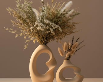 Puede incluir: Dos jarrones de cerámica beige con diseños únicos de círculo abierto, cada uno con arreglos florales secos. Los jarrones se colocan sobre una plataforma rectangular de piedra clara, creando una estética minimalista y terrosa. El jarrón más grande contiene un arreglo más completo.