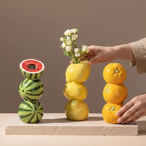 Puede incluir: Tres jarrones de cerámica con forma de fruta: una sandía, un limón con flores y una pila de naranjas. El jarrón de sandía es rojo y verde. El jarrón de limón contiene flores blancas. El jarrón de naranja es una pila de tres frutas de color naranja.