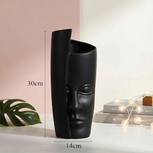 May include: A matte black ceramic vase featuring a sculpted human face on the front surface. The vase has an asymmetrical top edge and measures 30 cm in height and 14 cm in width, as indicated by the black dimension lines shown in the image.